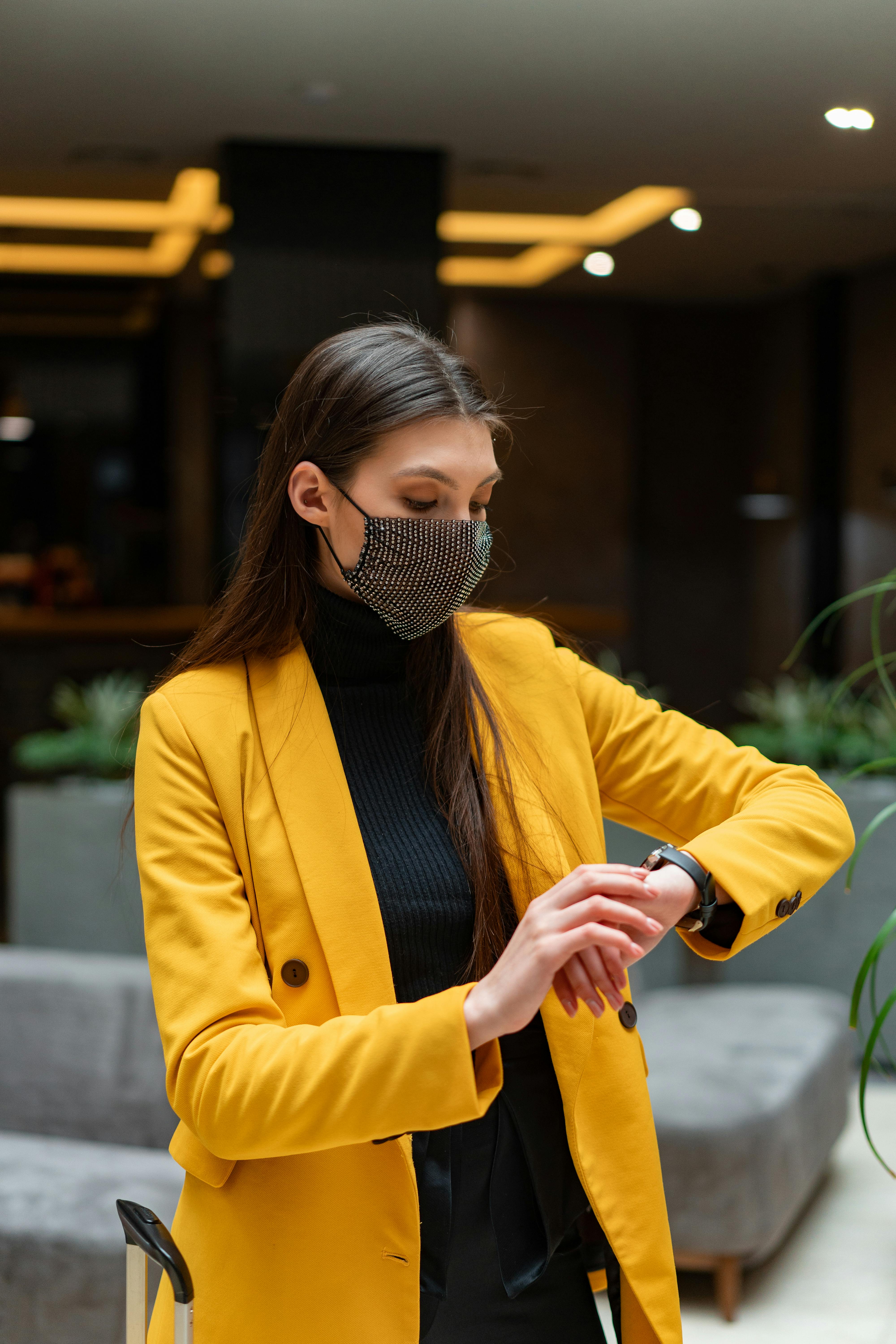 Fashionable woman wearing a face mask and yellow coat, checking time indoors.