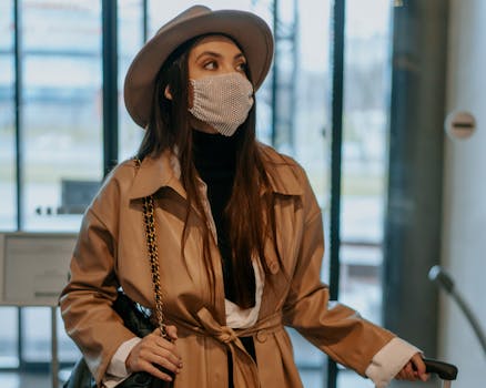 A fashionable woman in a brown coat and hat wearing a face mask inside an airport lobby.