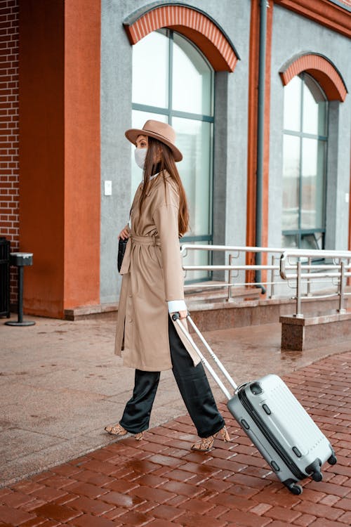 Photo of a Woman in a Brown Coat Walking with Her Gray Suitcase