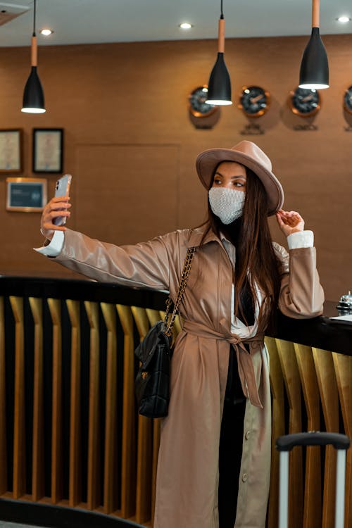 Stylish woman in brown hat and coat taking selfie in medical mask near wooden fence in modern apartment