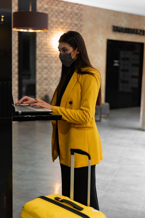 Free Woman wearing mask and yellow coat using laptop in hotel lobby next to suitcase. Stock Photo