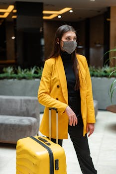 Fashionable woman wearing face mask and yellow blazer with suitcase at indoor airport.
