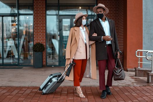 Fashionable couple with face masks at hotel entrance, showcasing travel during pandemic.