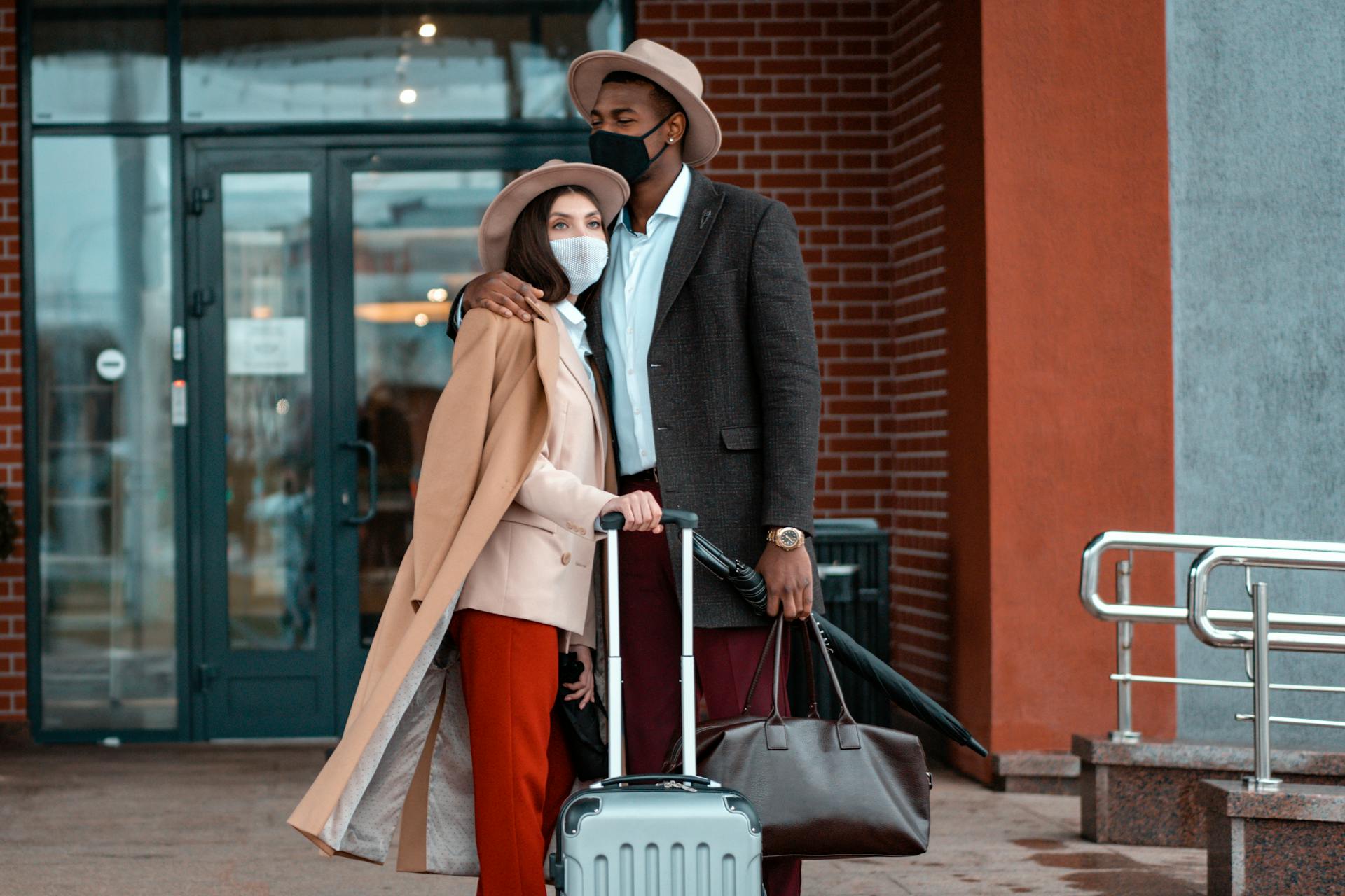 Couple wearing face masks standing with luggage outside modern building.