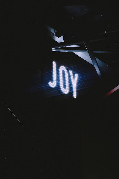 From below of illuminated signboard with bright neon light of Joy placed on dark background near metal structure