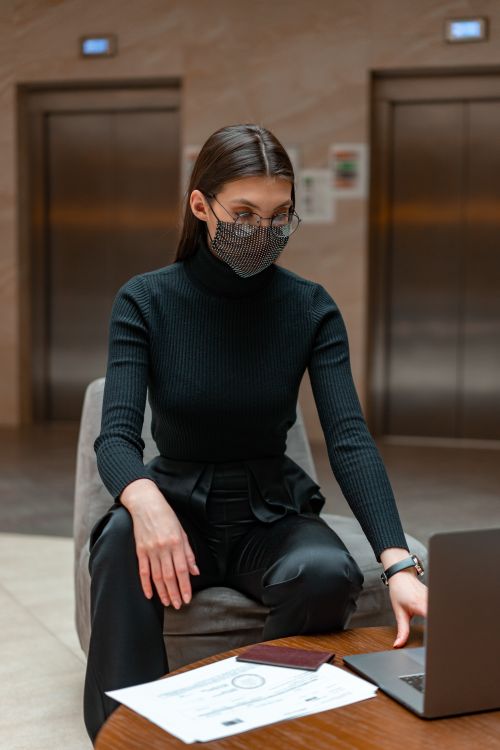 Free Stylish woman in black attire using laptop in a modern lobby setting, practicing safety with a mask. Stock Photo