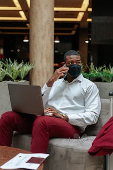 Businessman in mask using phone and laptop, sitting indoors.