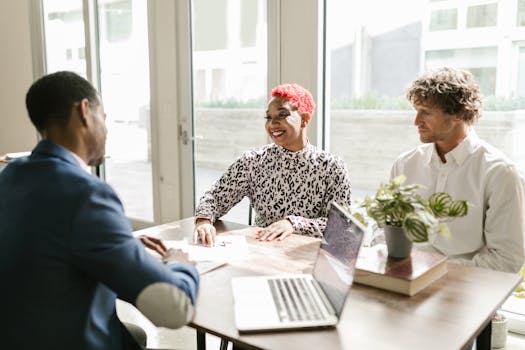 Three professionals meeting in a sunny office discussing finance with smiles.