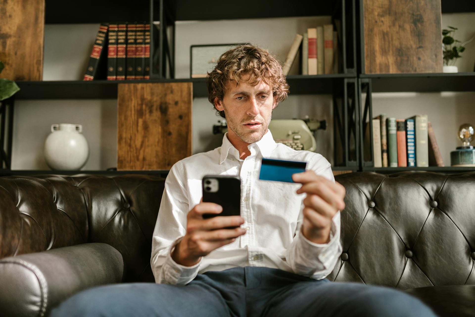 A man sitting on a leather sofa using a smartphone and credit card for online shopping.