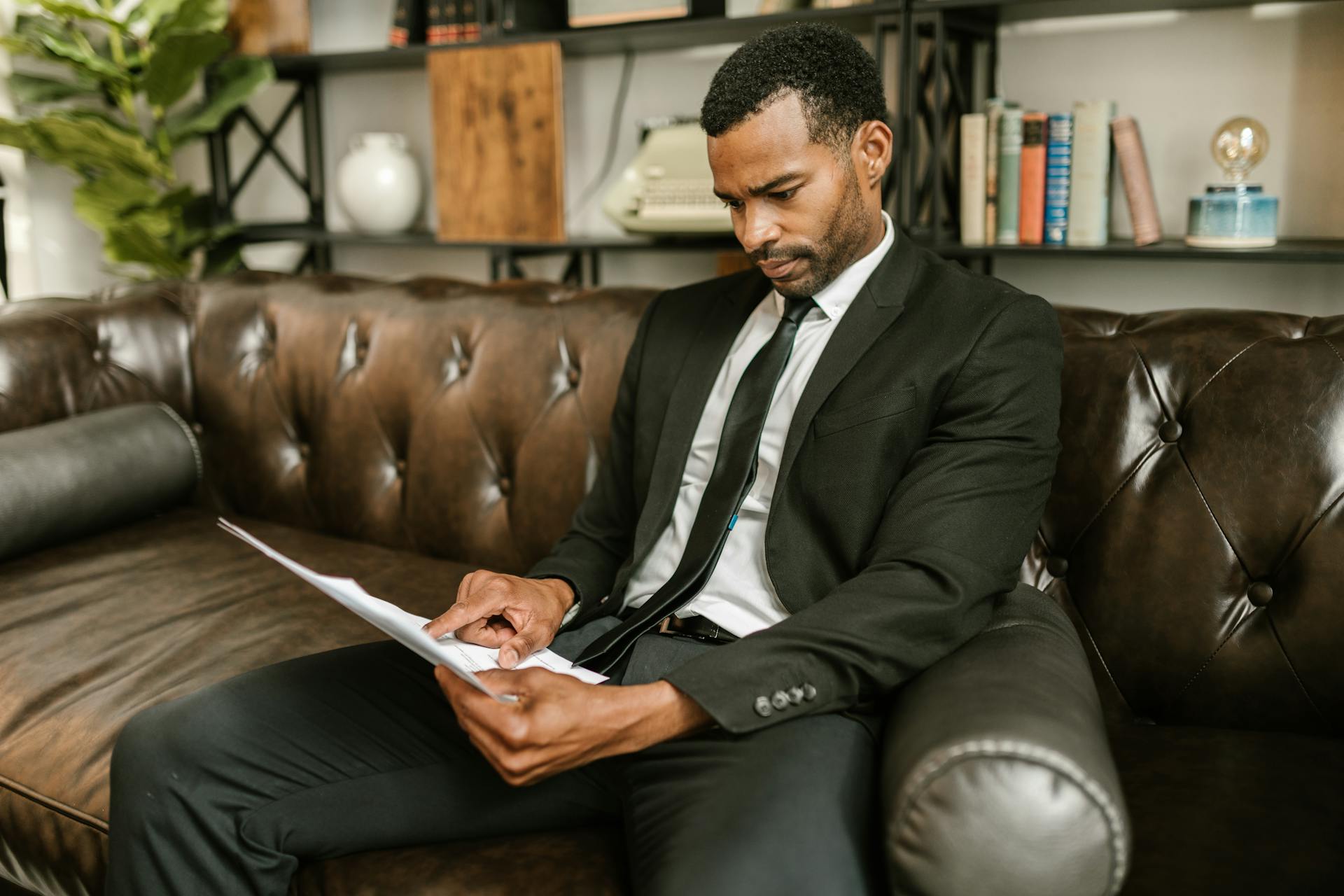 Businessman in corporate attire reading documents on a leather couch indoors.