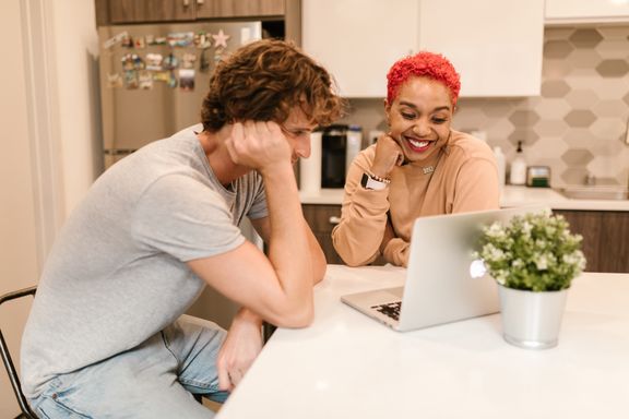 A couple leans on their kitchen counter smiling at an open laptop, a small plant beside it
