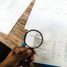 A person holds a magnifying glass over a printed financial document to closely inspect the account activity.