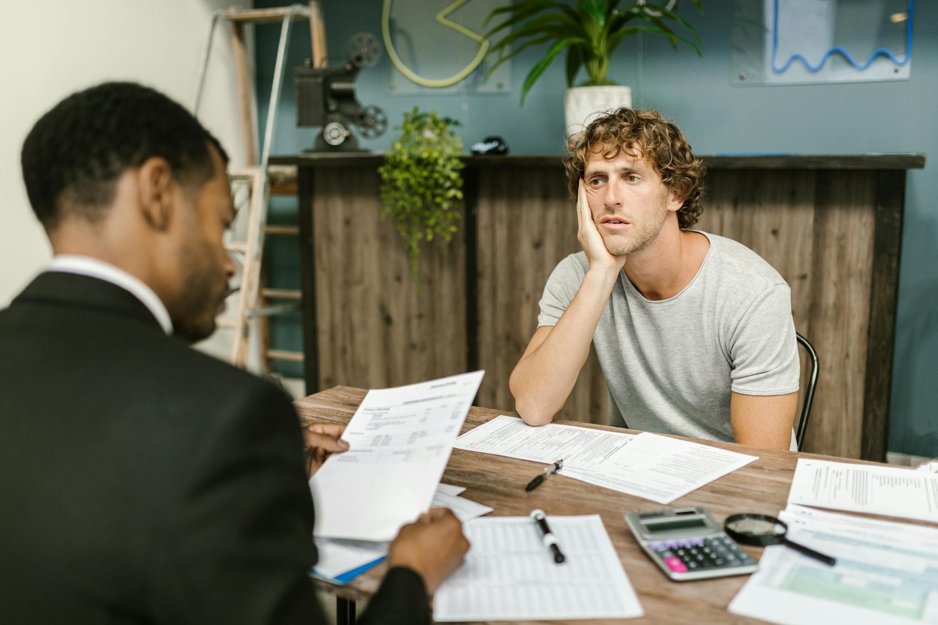 Man in office, appearing stressed during paperwork discussion, sitting opposite professional.