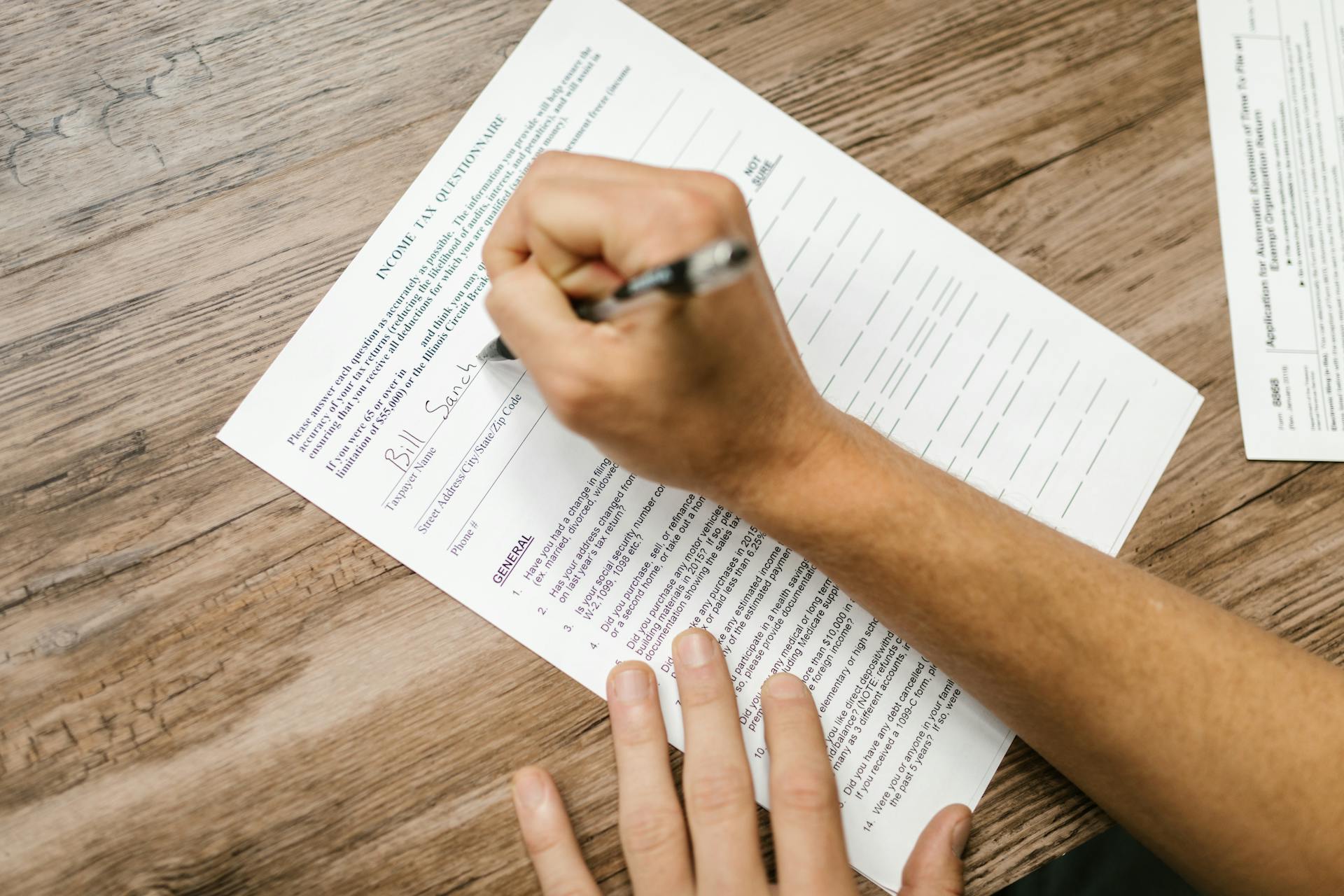 Person filling out a tax questionnaire with a pen, showing close-up on wooden table.