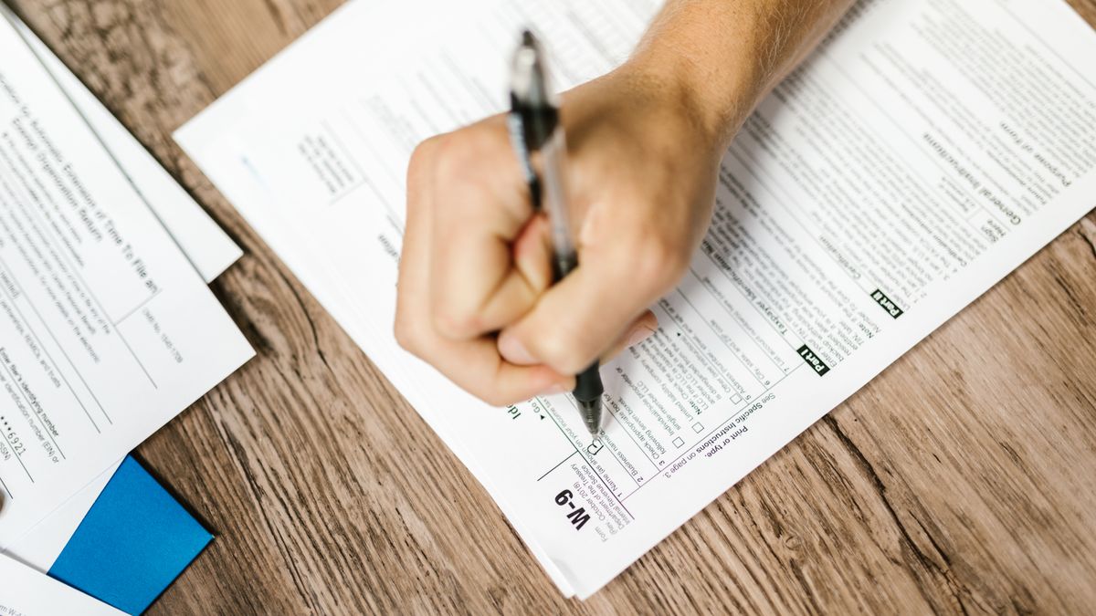 Hand writing on a W-9 tax form using a ballpoint pen on wooden desk.