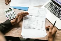 Manos sostienen documentos financieros con calculadora y laptop en un escritorio de oficina.