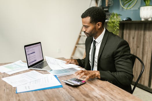 A businessman in a suit working on documents and using a laptop at an office desk.