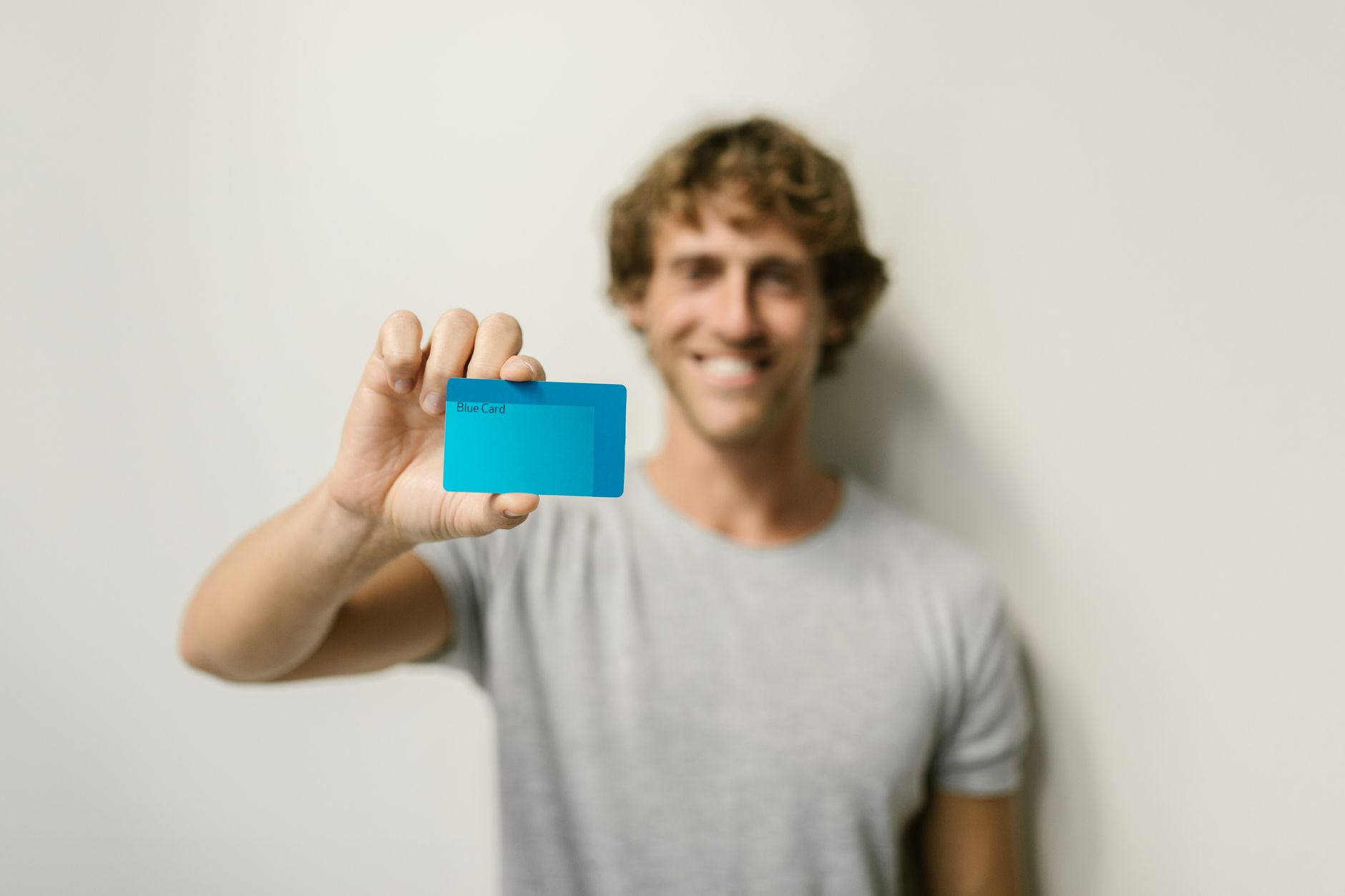 A smiling man in a gray shirt holds a blue card, offering a bright and friendly demeanor against a neutral backdrop.