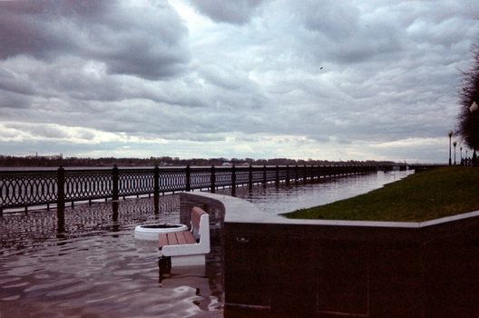 A flooded walkway by the river with overcast skies and empty benches submerged.