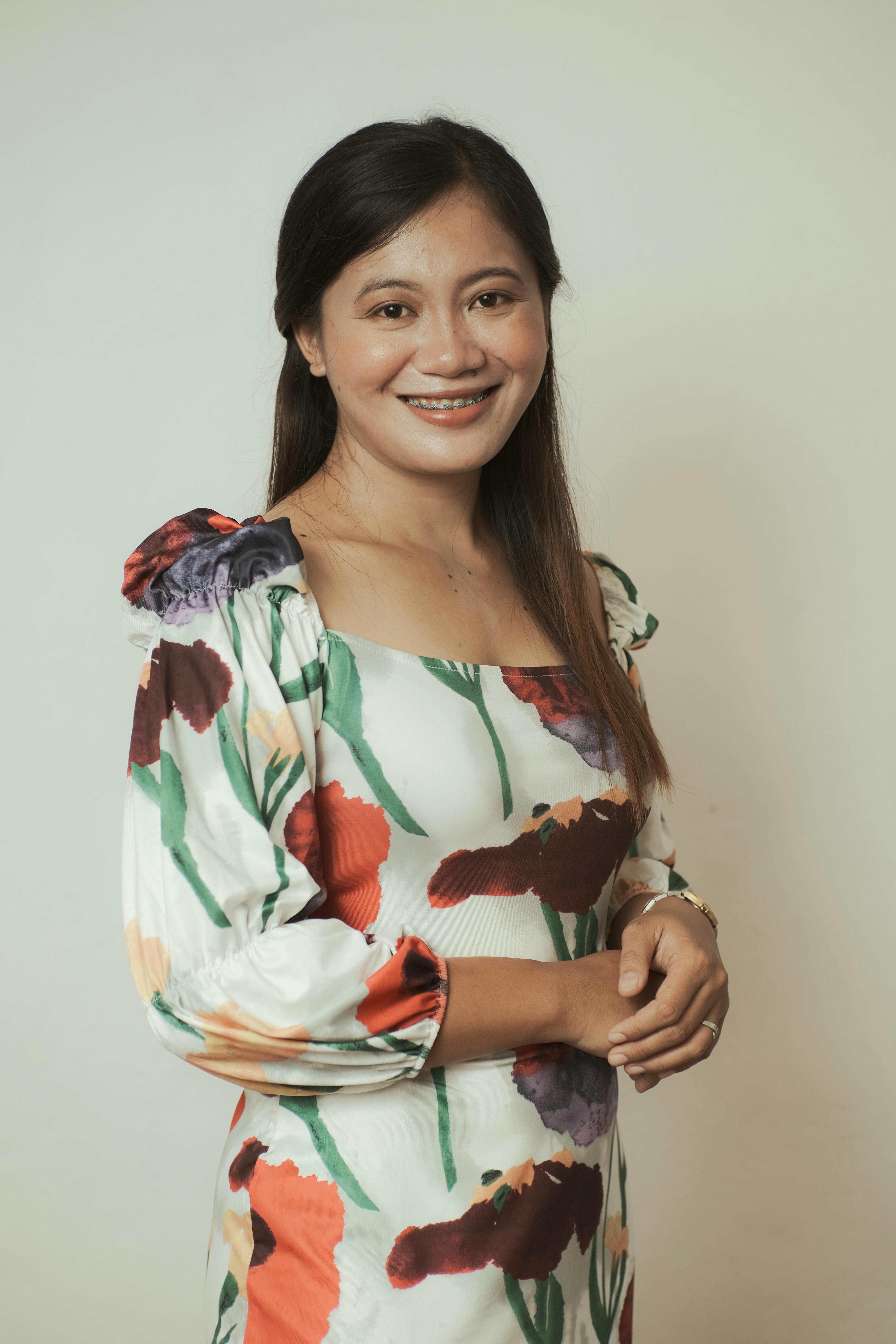 Portrait of an adult woman in a floral dress, smiling with hands clasped against a white background.