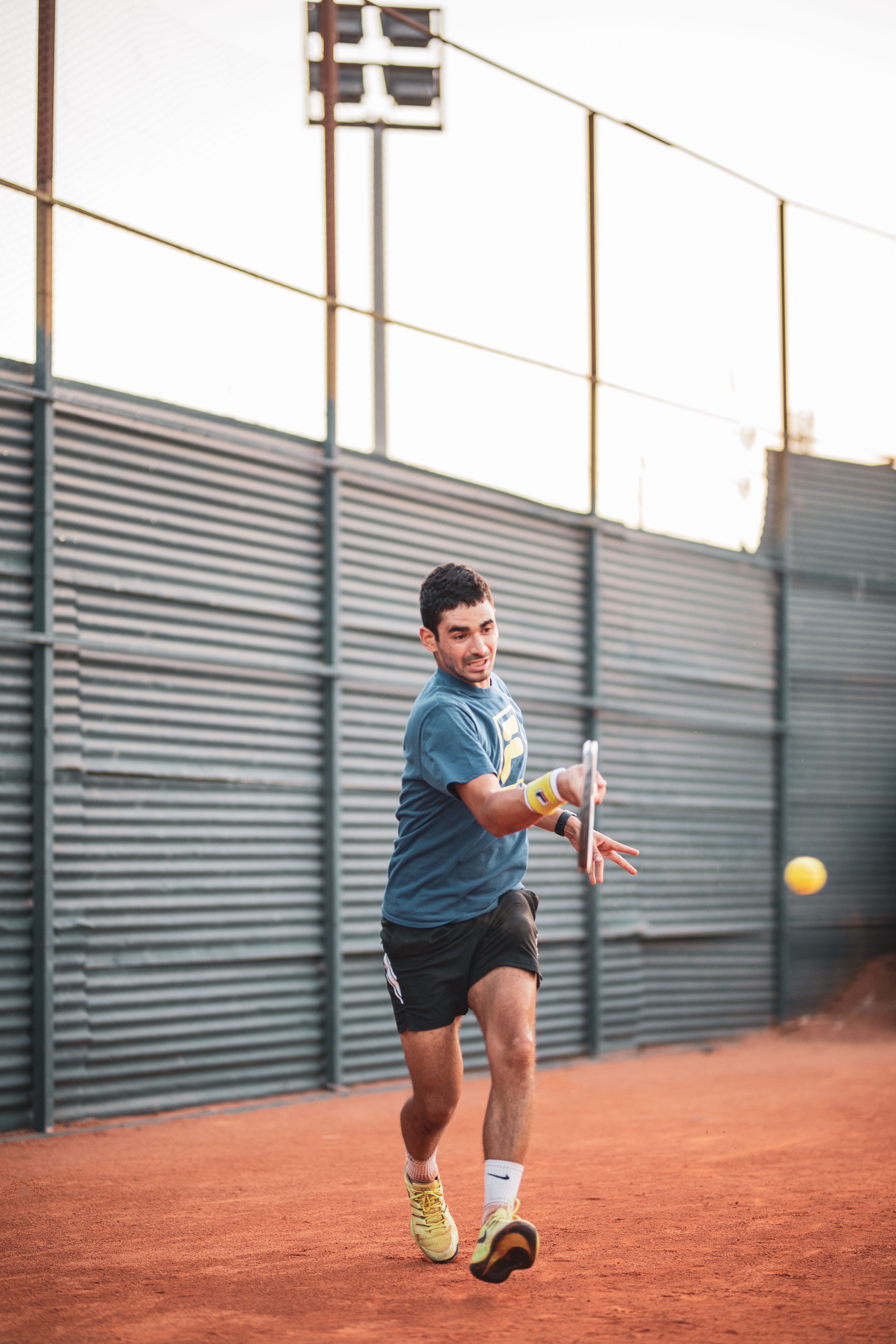 A young man energetically playing tennis on an outdoor clay court during the day.
