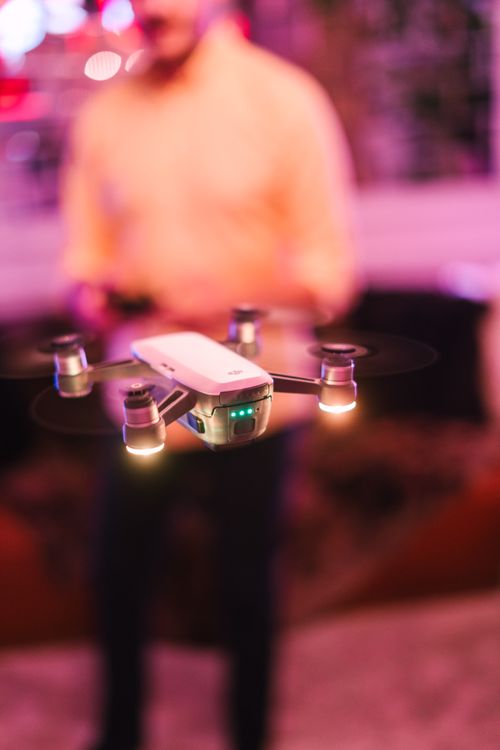 Free A mini drone hovers indoors with a blurred person in the background. Stock Photo