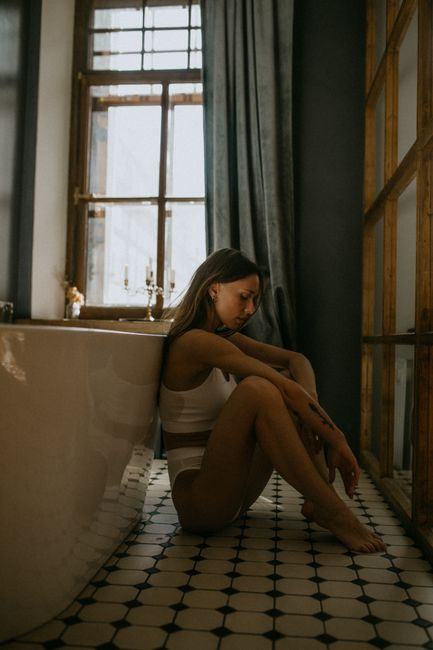 Side view of sensual female in underwear sitting on floor while leaning on bathtub on background of window