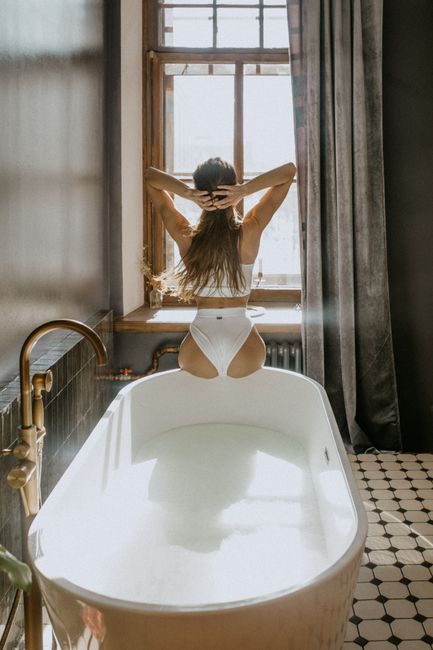 Back view of young female in swimwear sitting on bath edge with arms raised in front of window in daytime