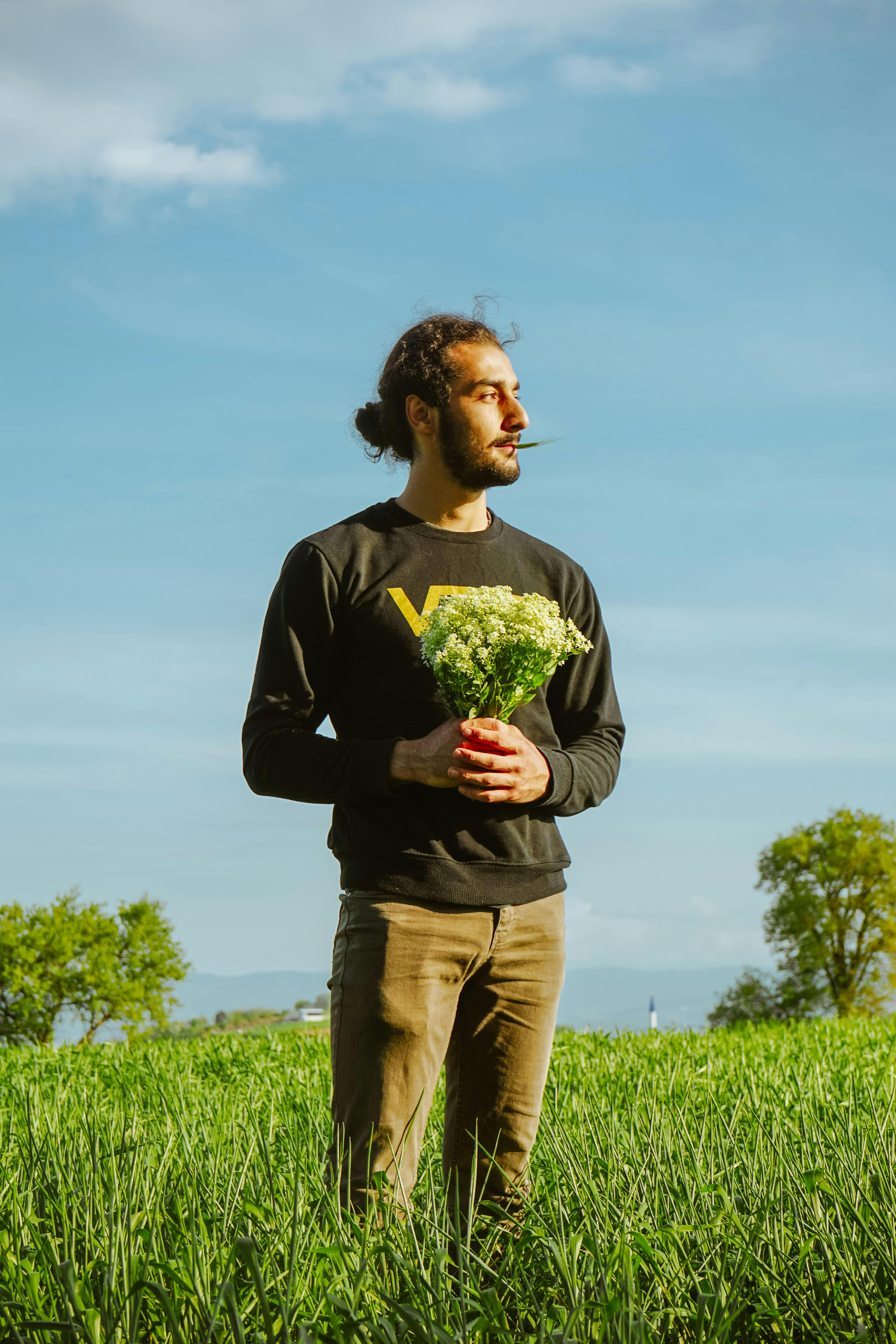 A bearded man stands in a lush field holding flowers under a clear blue sky.