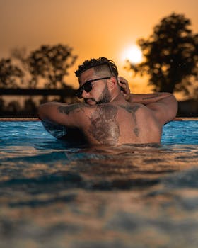 Shirtless man with tattoos and sunglasses enjoying a swim at sunset.