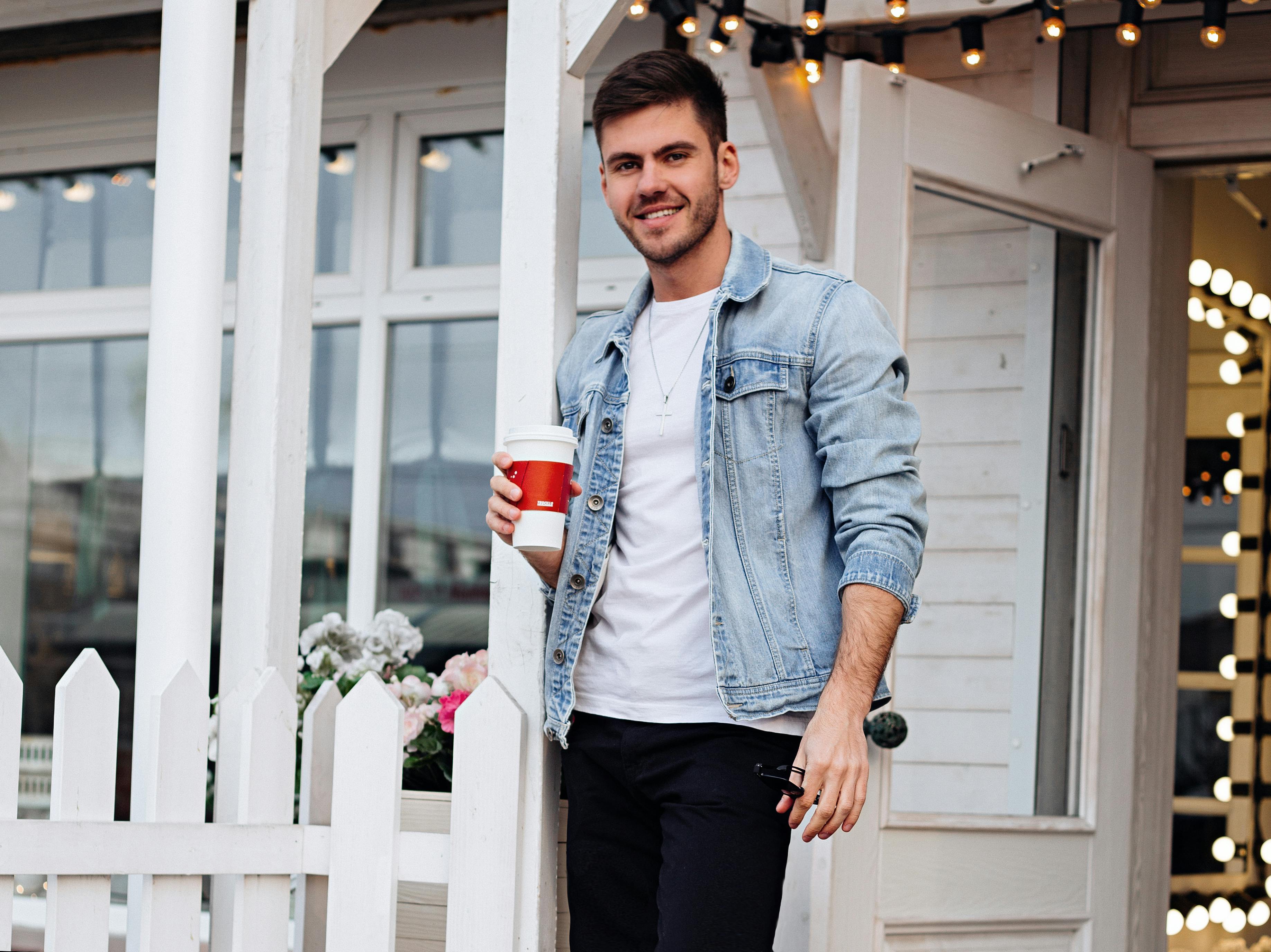 Stylish man in denim jacket smiling with coffee outside a cozy cafe.