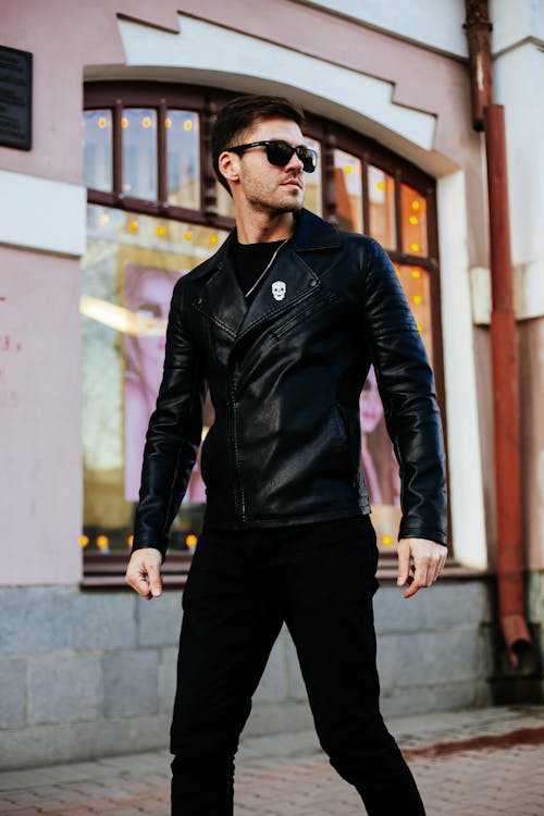 Man in Black Leather Jacket Wearing Sunglasses Standing Outside an Establishment while looking Afar