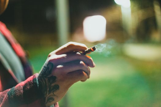 Close-up of a tattooed hand holding a lit joint outdoors at night with a blurred background.