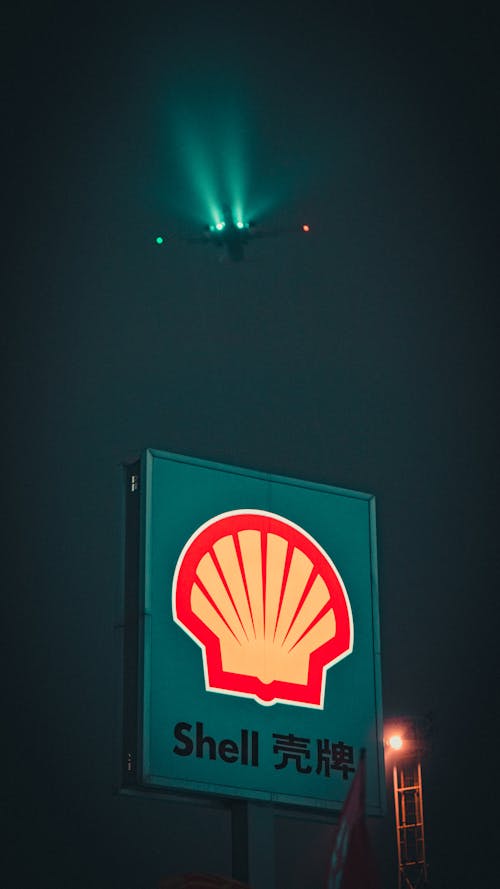 Banner with inscription and logotype of gas station located against dark sky with flying airplane at night time in city