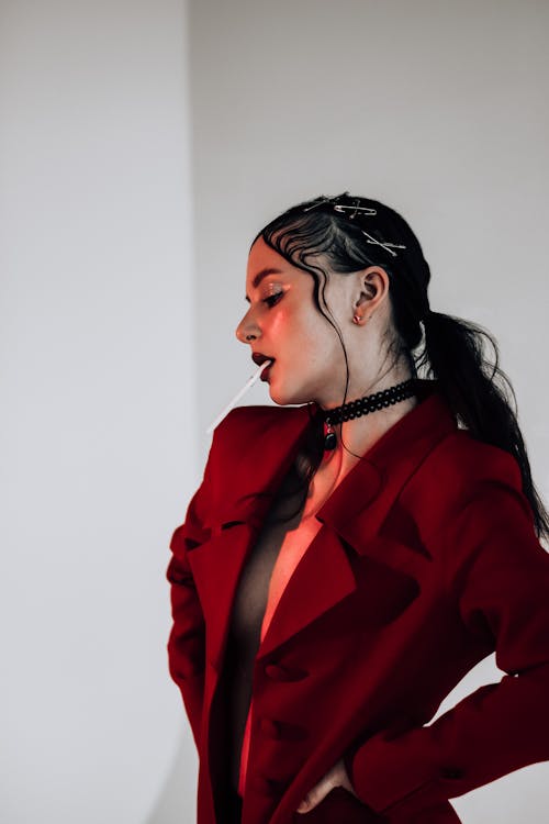 Serious young female in trendy red jacket with cigarette and makeup standing in red light in studio with closed eyes near white walls
