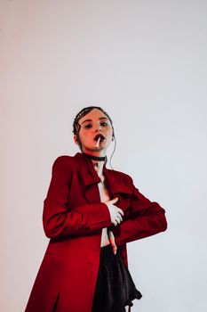 Confident young woman in trendy jacket with cigarette and makeup looking at camera while standing in bright room with white wall