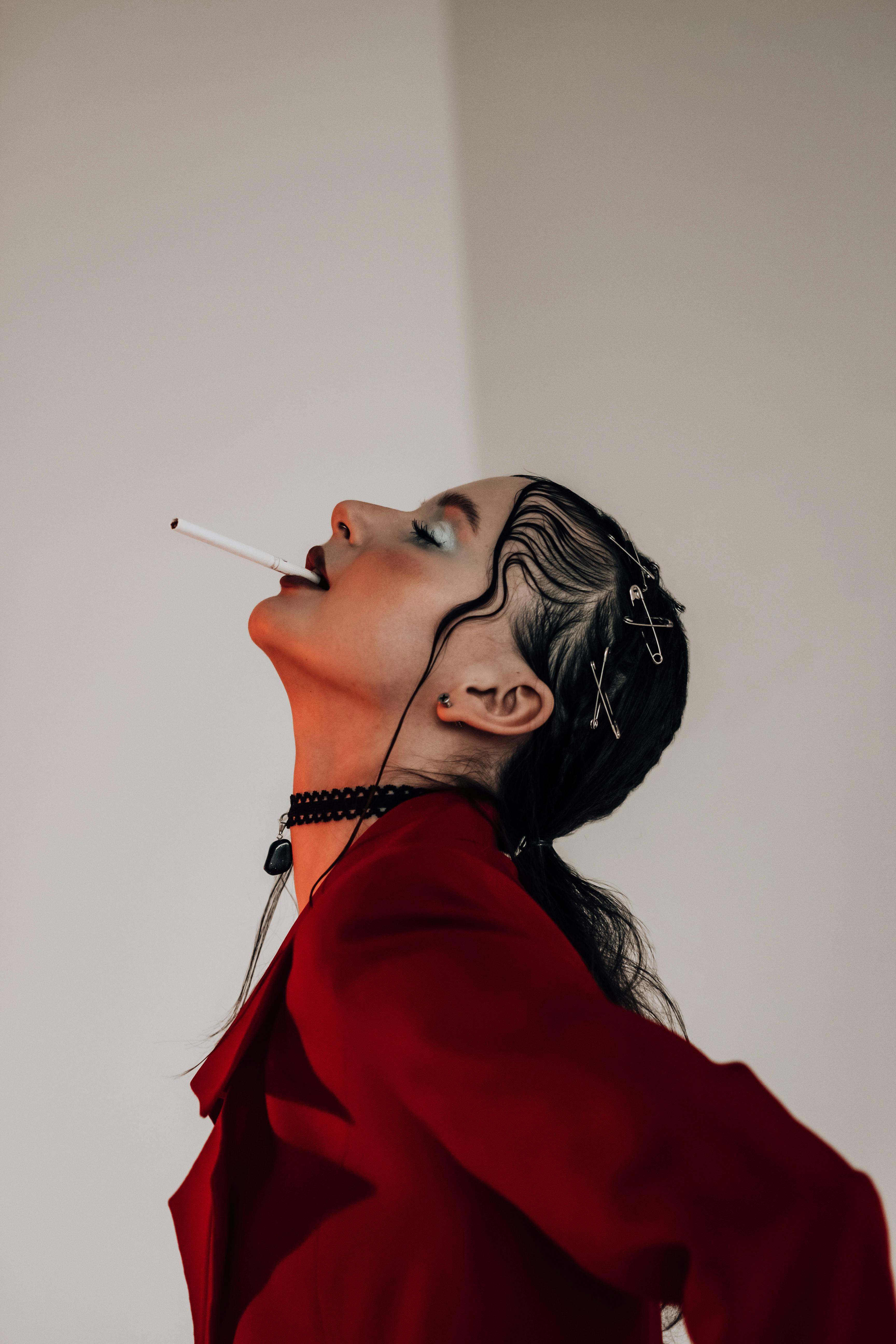 Side view of thoughtful young female in trendy red jacket and makeup with cigarette in lips and closed eyes in red light in studio on white background