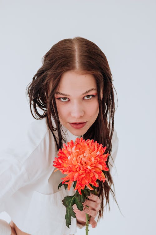 Positive young woman in stylish clothes with red chrysanthemum looking at camera on white background in bright studio