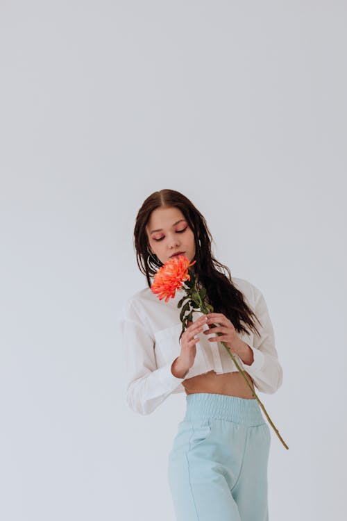 Pensive young woman in stylish clothes with red chrysanthemum in hand with closed eyes on white background in bright studio