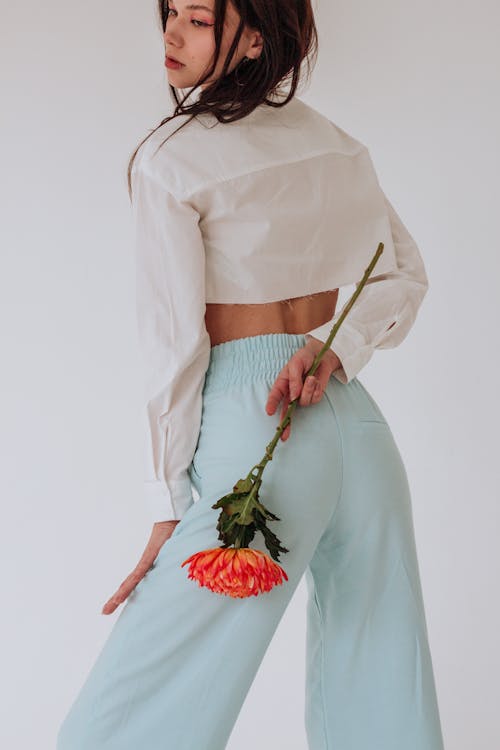 Back view of pensive young woman in stylish clothes with red chrysanthemum in hand behind back looking away on white background in bright studio