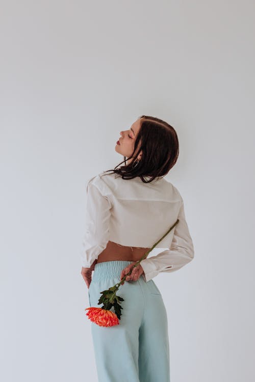 Back view of thoughtful young woman in stylish clothes standing with red chrysanthemum in hand behind back with closed eyes on white background in bright studio