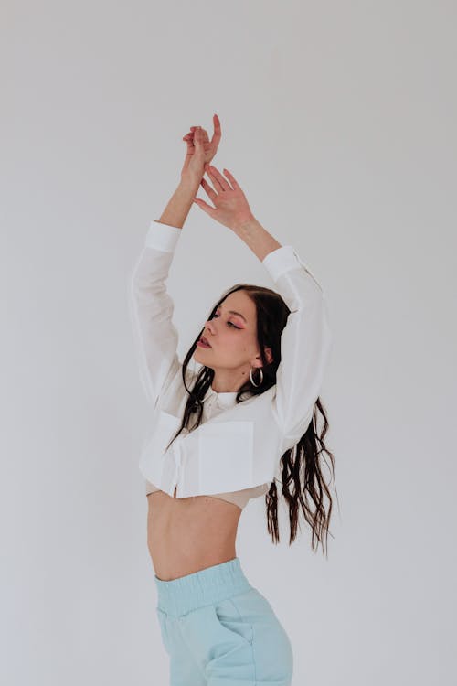 Free Thoughtful young female in stylish clothes standing with raised hands and closed eyes on white background in bright studio Stock Photo