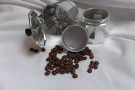 Still life of a disassembled moka pot with scattered roasted coffee beans on a white surface.