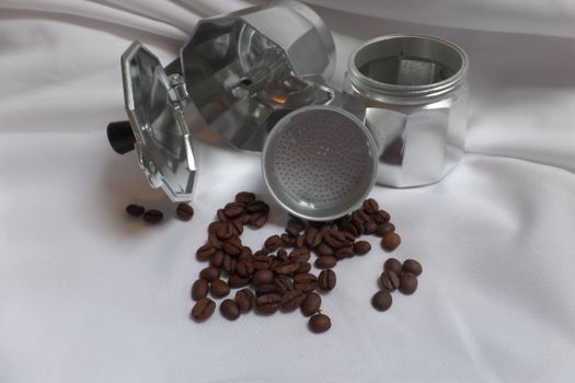 Still life of a disassembled moka pot with scattered roasted coffee beans on a white surface.