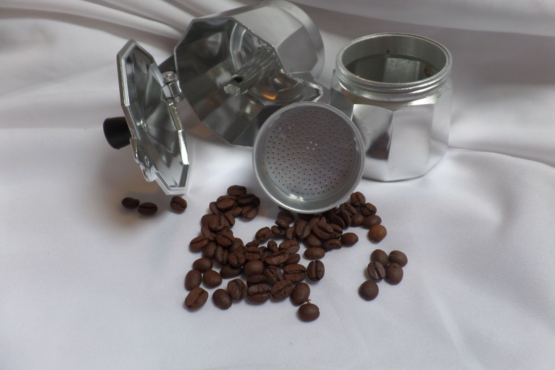 Still life of a disassembled moka pot with scattered roasted coffee beans on a white surface.
