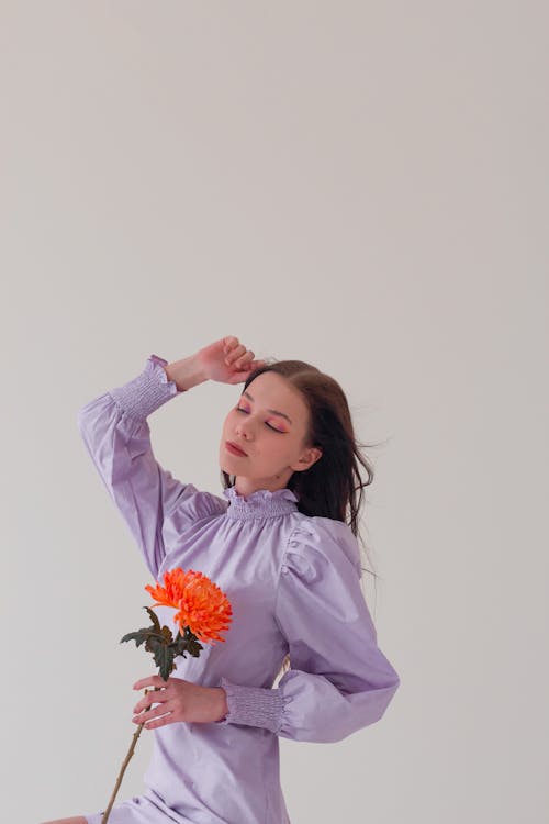 Charming female in elegant dress holding flower while touching hair with closed eyes in studio with white wall