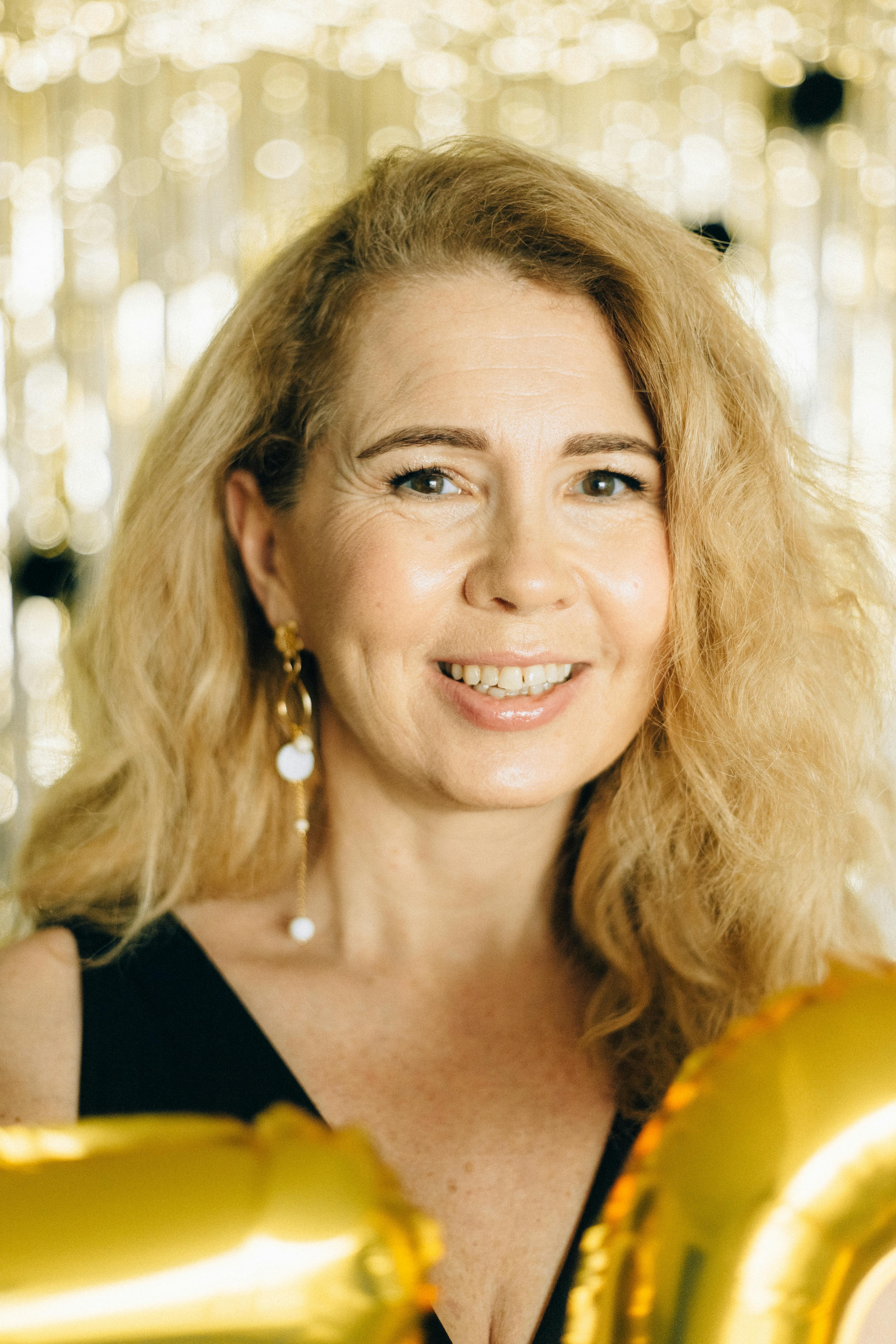 Smiling senior woman with blonde hair in elegant attire holding golden balloons against a shimmering backdrop.