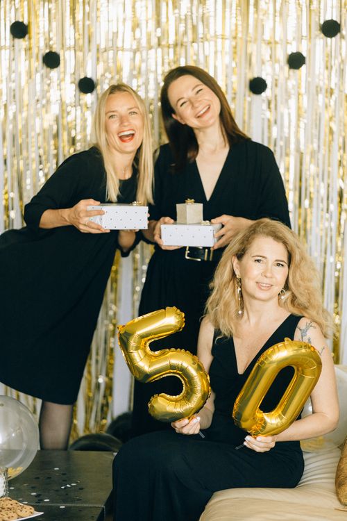 Free Three women celebrating a 50th birthday with gifts and golden balloons indoors. Stock Photo