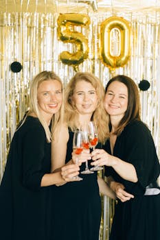 Three women celebrating a 50th birthday with champagne, wearing black dresses and smiling.