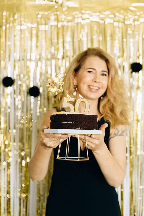 Free Happy woman holding 50th birthday cake with golden decorations indoors. Stock Photo